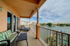 A balcony with a sofa, chairs, and a table overlooking a body of water with boats and buildings in the background.