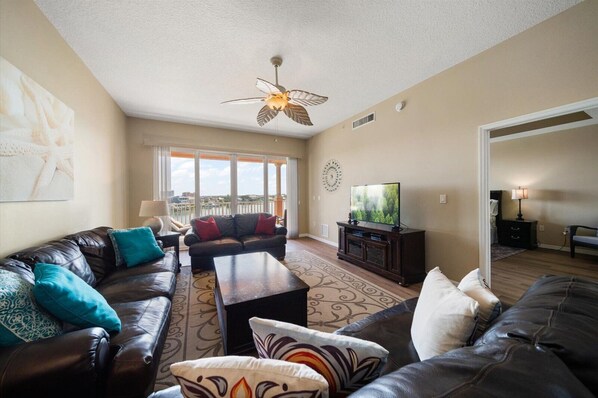 A spacious living room with a large sofa set, a coffee table, and a television. There is a ceiling fan and a view of the outdoors through large windows.