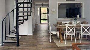 Un interior moderno con una escalera de caracol, una zona de comedor con sillas blancas y un ventilador de techo.
