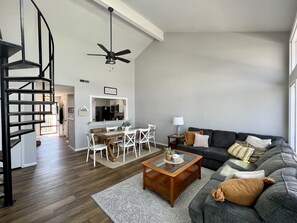 A modern living room with a dark gray sofa, a wooden coffee table, and a dining area with white chairs. A black ceiling fan is visible, and a staircase leads to another level.