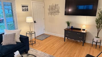 A living room with a dark blue sofa, a flat-screen TV, a small side table, a potted plant, and a wooden floor.