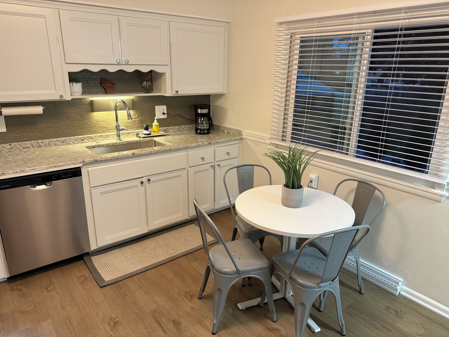 A kitchen with white cabinets, a stainless steel dishwasher, a granite countertop, a coffee maker, and a potted plant on a round table with two chairs.