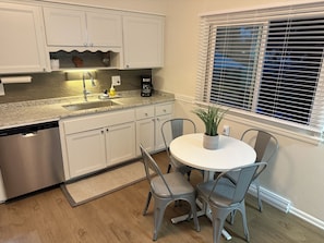 Una cocina con gabinetes blancos, lavavajillas de acero inoxidable, encimera de granito, cafetera y planta en maceta sobre una mesa redonda con dos sillas.