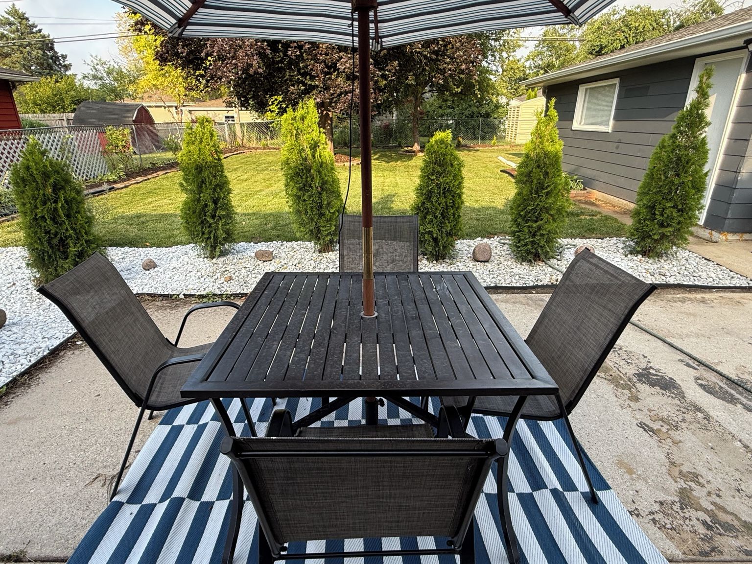 An outdoor patio with a table and chairs, surrounded by a garden with trimmed bushes and a lawn.