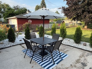Un patio con una mesa y sillas, un paraguas a rayas, y un pequeño jardín con arbustos y una valla.