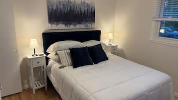A bedroom with a bed, two bedside tables, a nightstand, a window with blinds, and a painting on the wall.