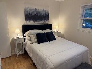 Un dormitorio con cama, dos mesitas de noche, una mesilla auxiliar, una ventana con persianas y un cuadro en la pared.