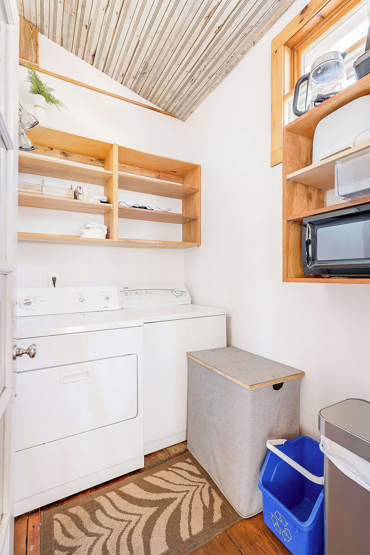 A laundry room with a white washer and dryer, wooden shelves, a blue recycling bin, and a grey storage box.