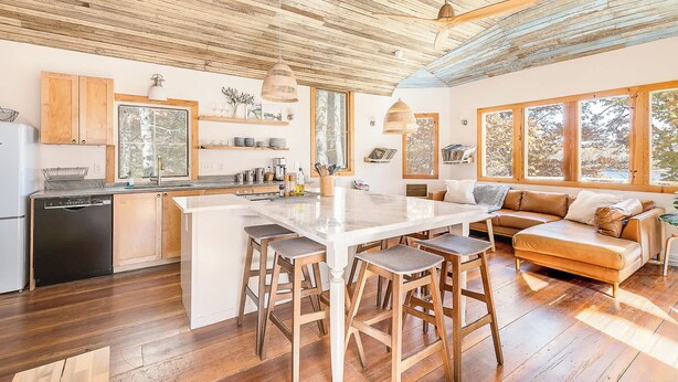 A spacious kitchen with wooden cabinets, a large island, and bar stools. There is a dining area with a sofa and a large window providing a view of the outdoors. - The Lake House: Dock|Marine Hammock|Screen Porch (Prospect Hill)