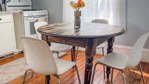 A dining area with a round table, white chairs, a vase with flowers, a kitchen with white cabinets and appliances, and a window with sheer curtains. - Studio with Deck, Walk to Duke (Durham)