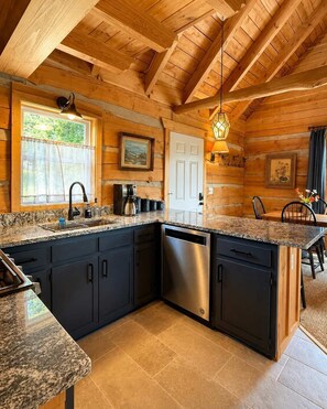 A kitchen with granite countertops, dark cabinets, a dishwasher, and a window with a view of greenery. - Scenic Log Cabin in Meadows of Dan (Meadows of Dan)