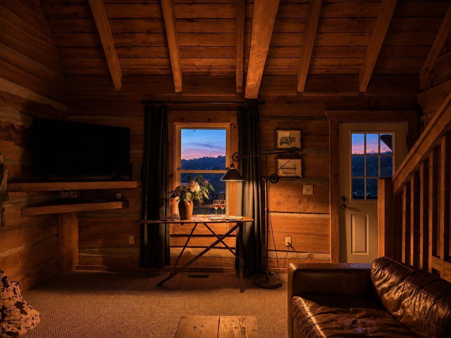 A cozy cabin interior with a wooden ceiling, a television mounted on the wall, a window with a view, a leather sofa, and a small table with a vase and glasses.