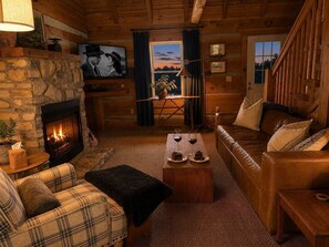 A cozy log cabin interior with a stone fireplace, a plaid sofa, a wooden coffee table, and a view through a door. - Scenic Log Cabin in Meadows of Dan (Meadows of Dan)