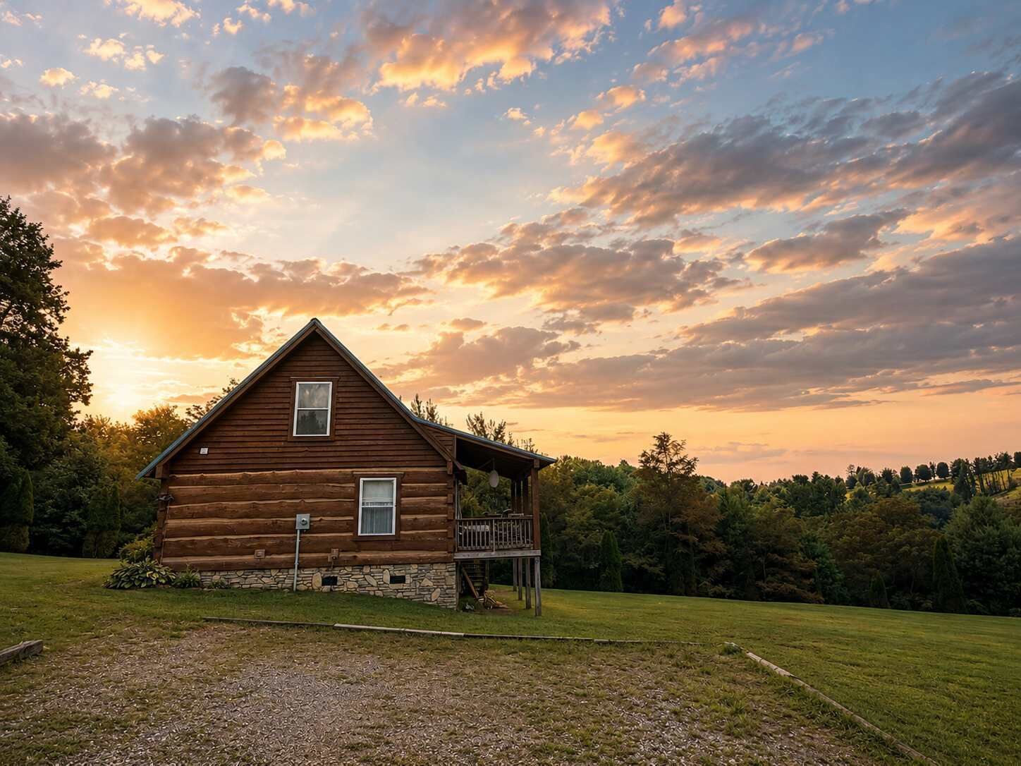 A wooden cabin with a porch, surrounded by a grassy field and trees, under a sunset sky.