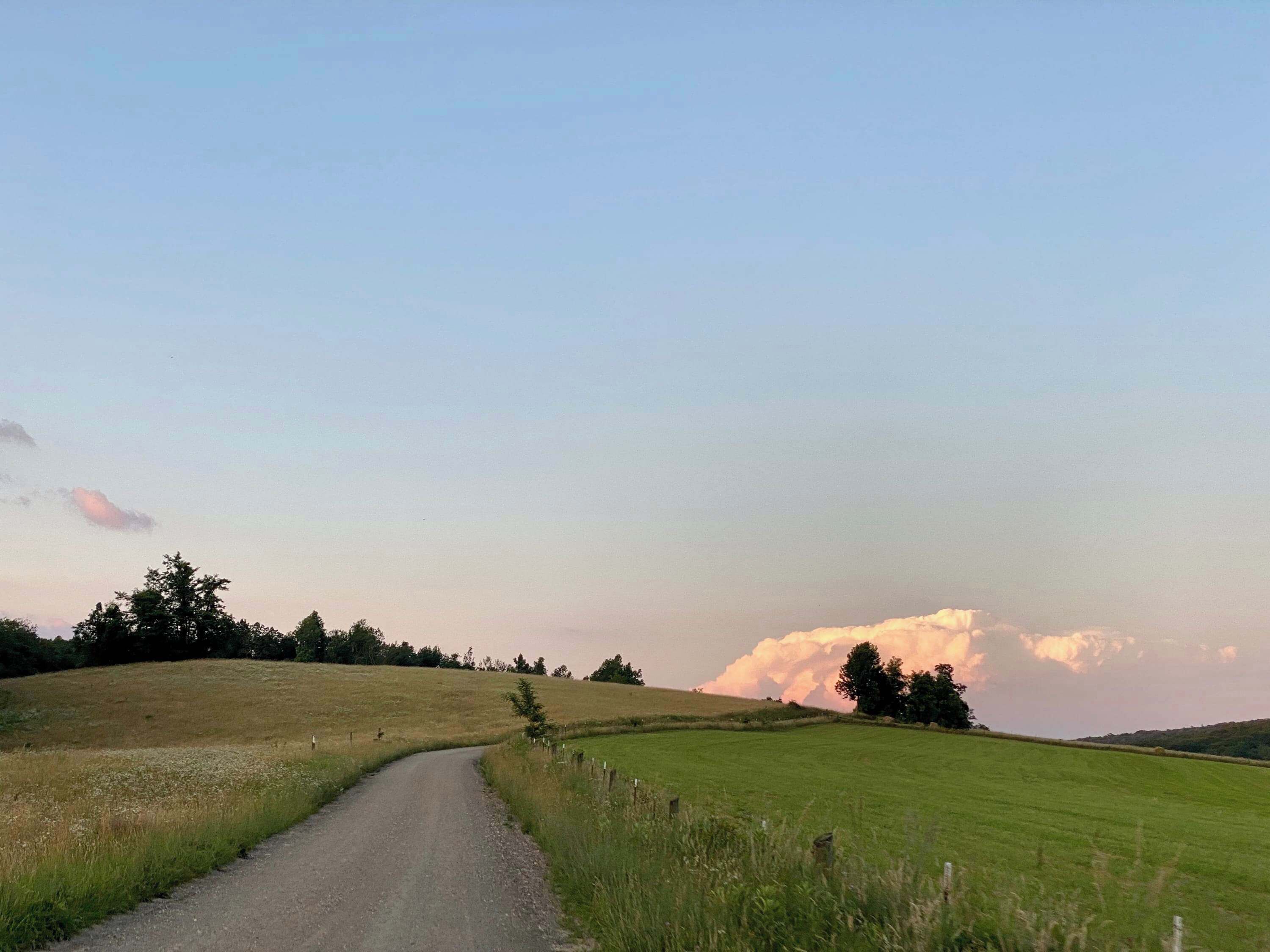 A rural road curving through a green field with a large cloud in the sky.