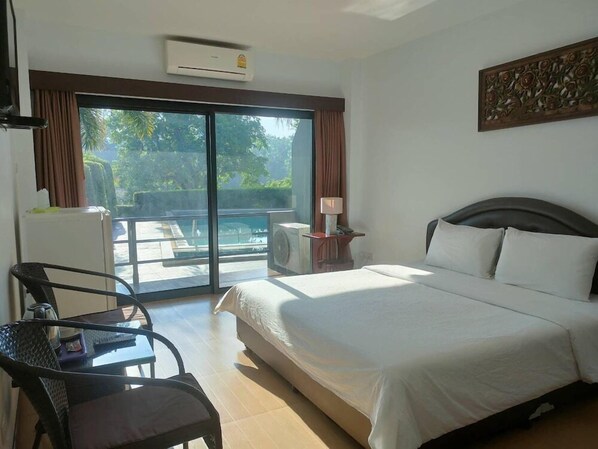 A hotel room with a bed, a chair, a table, an air conditioning unit, and a view of a pool and greenery. - Evergreen Resort Chanthaburi (Makham)
