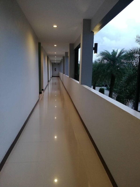 Un corredor largo y bien iluminado con piso reflectante, que conduce a un balcón con vistas a palmeras.