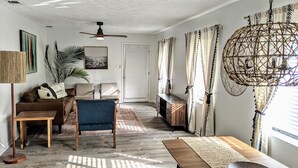 A living room with a sofa, chairs, a coffee table, a floor lamp, a ceiling fan, and a large window with patterned curtains.