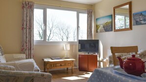 Een knus ogende kamer met een bed, een televisie, een tafel met theepot en een raam met gordijnen.
