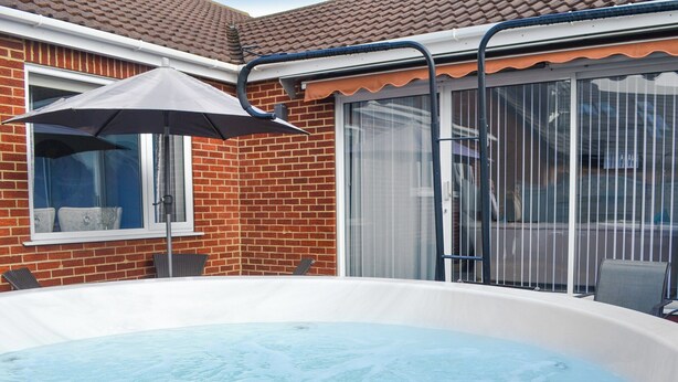 A hot tub in a backyard with a brick house, a window, and a patio chair.