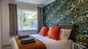 A bedroom with a large bed, a window with curtains, a tropical wallpaper, and a bedside table.