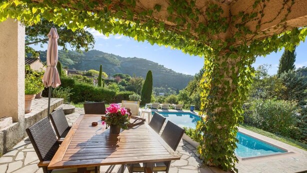 Ein Holztisch mit Stühlen, ein rosa Sonnenschirm und eine Blumenvase in einem überdachten Außenbereich mit Pool und Grünflächen im Hintergrund.