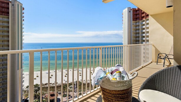 Un balcon avec un panier en osier contenant des serviettes et une chaise, donnant sur une plage avec des bâtiments en arrière-plan.