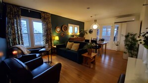 A living room with a dark green wall, a wooden floor, a sofa, a coffee table, a dining table, and a laptop on a small table.