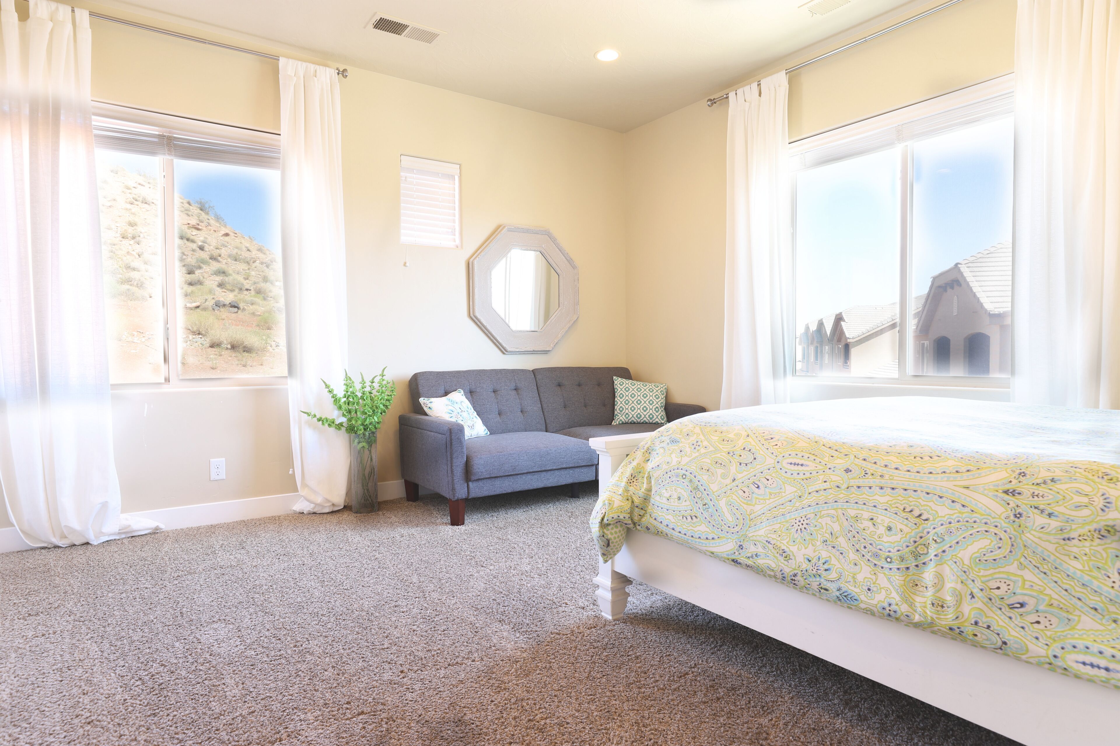 A bedroom with a bed, a grey sofa, a large window with a view of a hill, and a mirror on the wall.