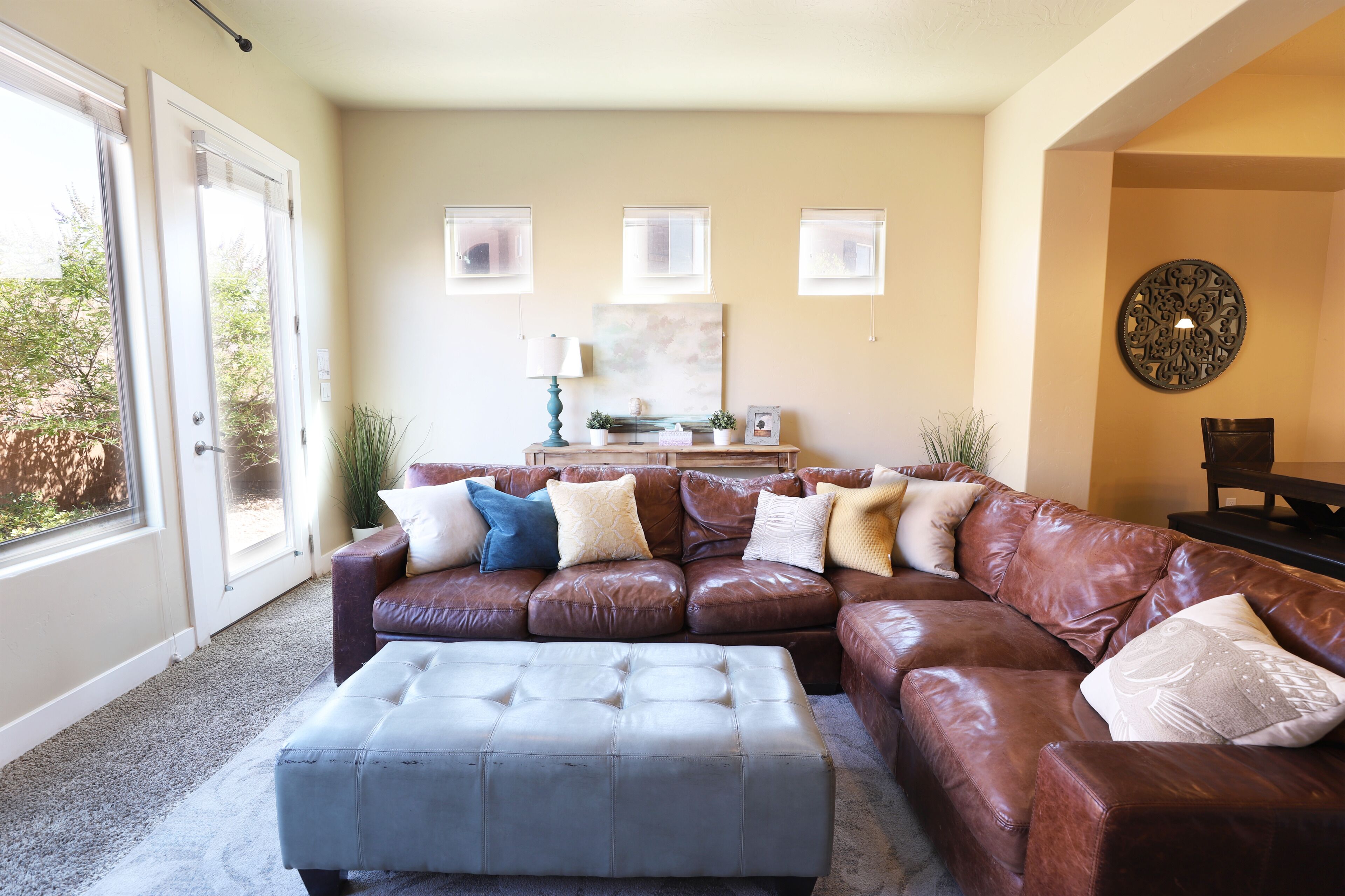 A living room with a brown leather sectional sofa, a grey ottoman, and a coffee table. There are three windows with blinds, a painting on the wall, and a decorative wall piece.