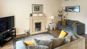A living room with a fireplace, a television, a grey sofa with yellow cushions, and a framed picture above the fireplace. - 2 bedroom accommodation in Bridlington (Bridlington)