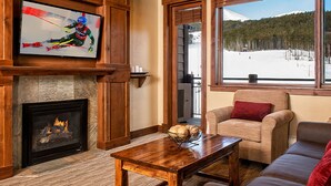 Un salon chaleureux avec une cheminée, un téléviseur fixé à un mur en bois, un canapé confortable et une grande fenêtre donnant sur un paysage enneigé.