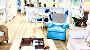 Un vaste espace de vie comprenant une table basse blanche, un fauteuil bleu, un canapé blanc et, au fond, une cuisine.