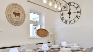 A dining area with a large wall clock, a hanging light fixture, and a rustic wooden table set for a meal. - 4 bedroom accommodation in West Bennan, near Shannochie (West Bennan, near Shannochie)