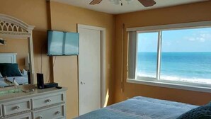 Een slaapkamer met een bed, een plafondventilator, een televisie, een dressoir en een raam met uitzicht op de oceaan.