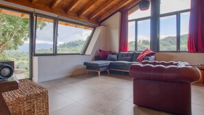 A modern living room with a wooden ceiling, large windows with red curtains, a grey sectional sofa, a red ottoman, and a wicker basket.