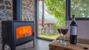 A wood-burning stove, a bottle of wine, and two glasses on a wooden table in a room with stone walls and a view of the outdoors.