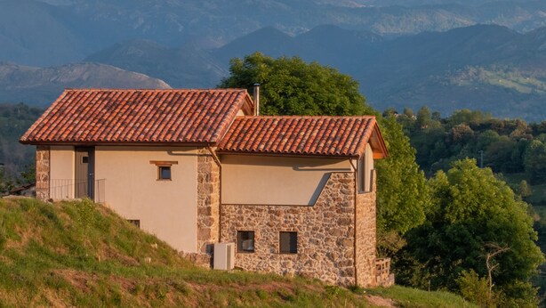 A stone house with a tiled roof, set against a mountainous backdrop.