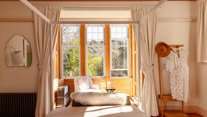 A bedroom with a large window, a bed, a chair, a radiator, and a wooden floor. - Home Farm (Oban)