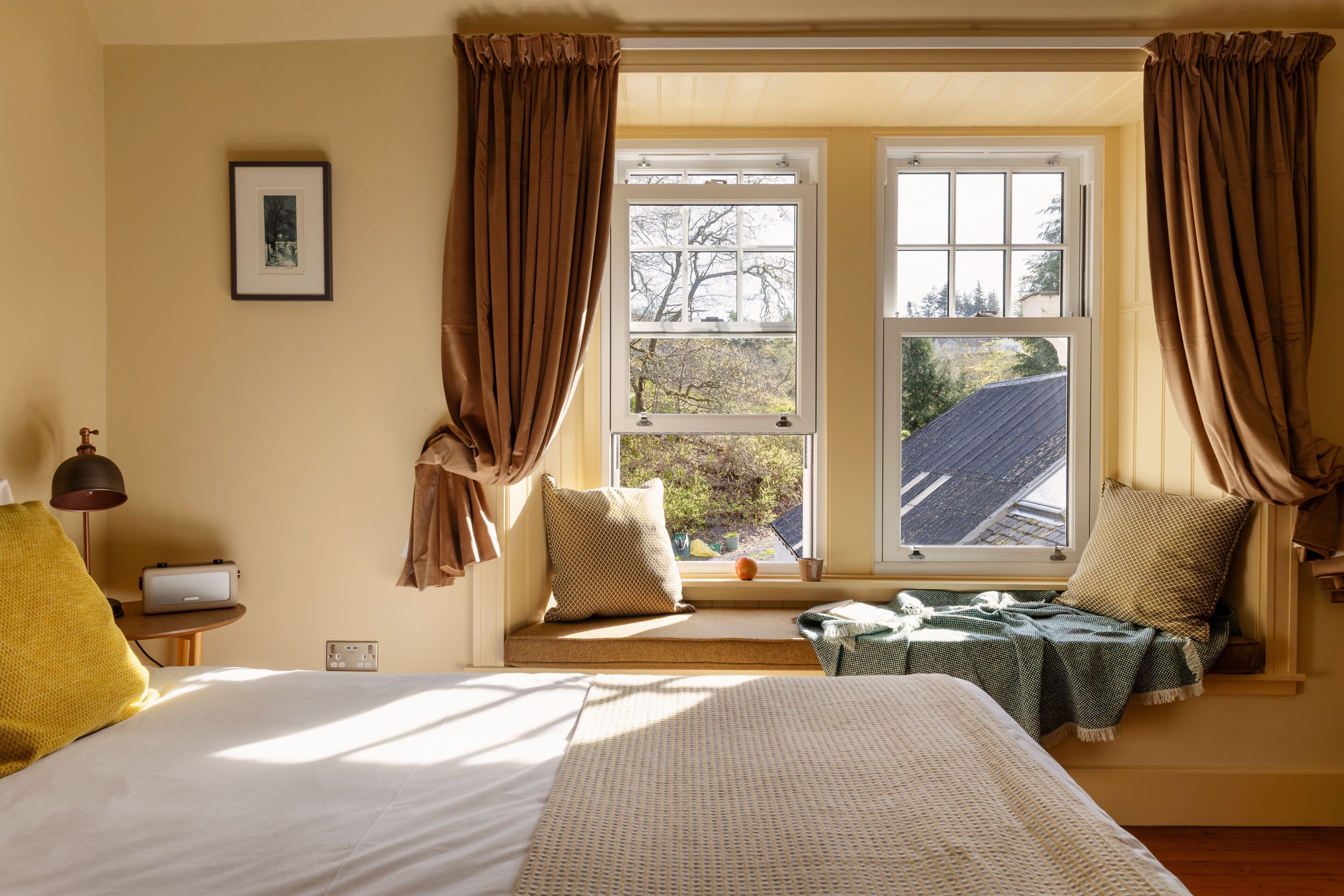 A bedroom with a large bed, a window seat with a view, and a bedside table with a lamp.