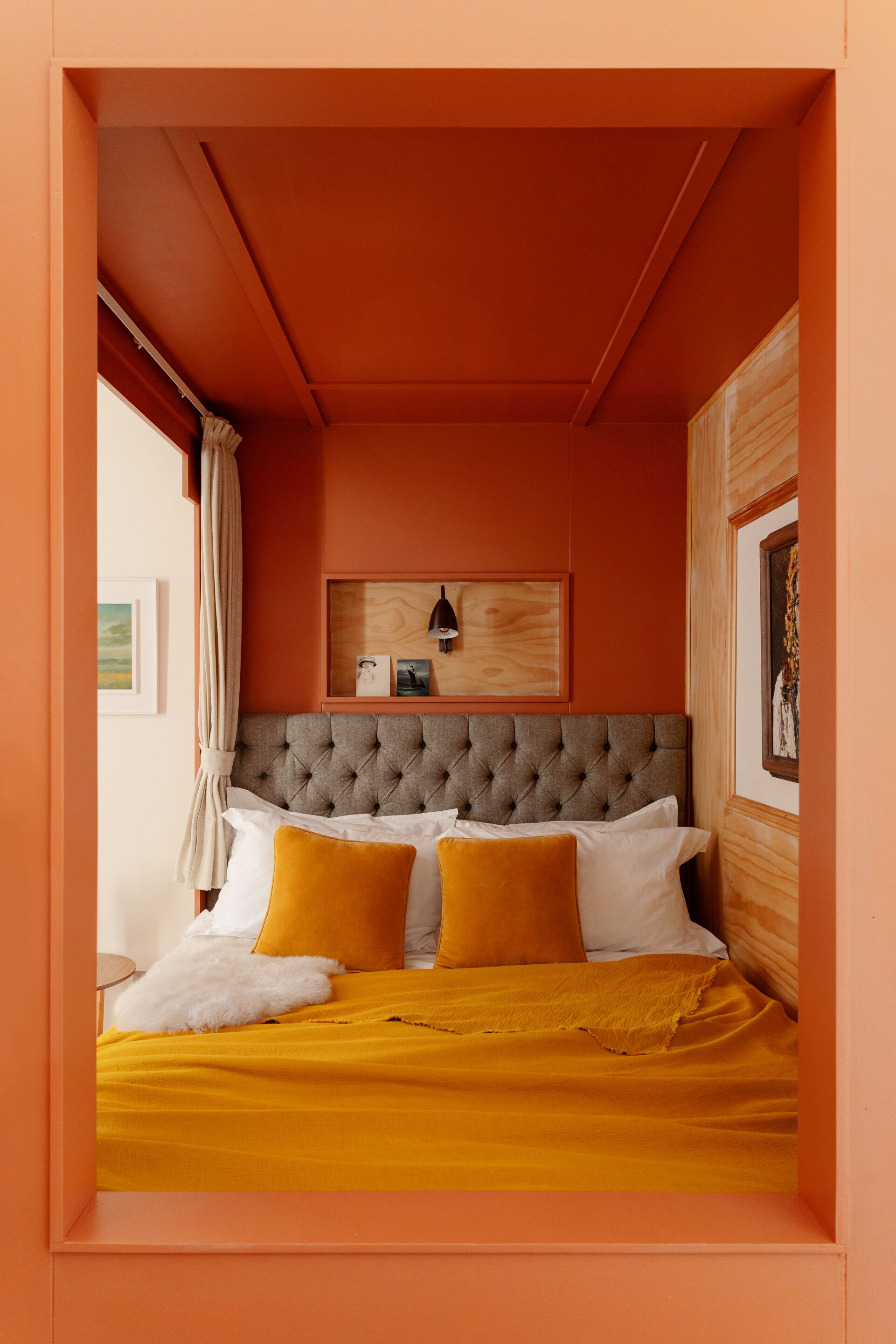 A bedroom with a tufted headboard, a bed with yellow and white bedding, a wooden shelf with decorative items, and a window with curtains.