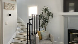 Un escalier avec des rampes noires, un fauteuil gris avec un coussin jaune, une plante en pot et un téléviseur à écran plat fixé au-dessus d’une cheminée.