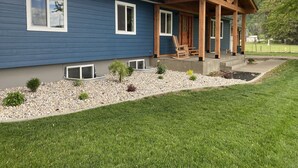 Uma casa moderna com fachada azul, varanda coberta com vigas de madeira e um pequeno jardim com pedras e plantas.