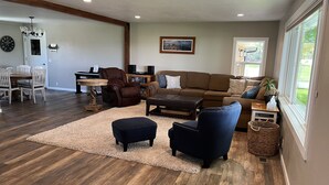 Uma sala de estar espaçosa com um sofá de couro marrom, uma poltrona azul e uma mesa de centro. Há uma área de jantar com mesa e cadeiras de madeira, e um piano ao fundo.