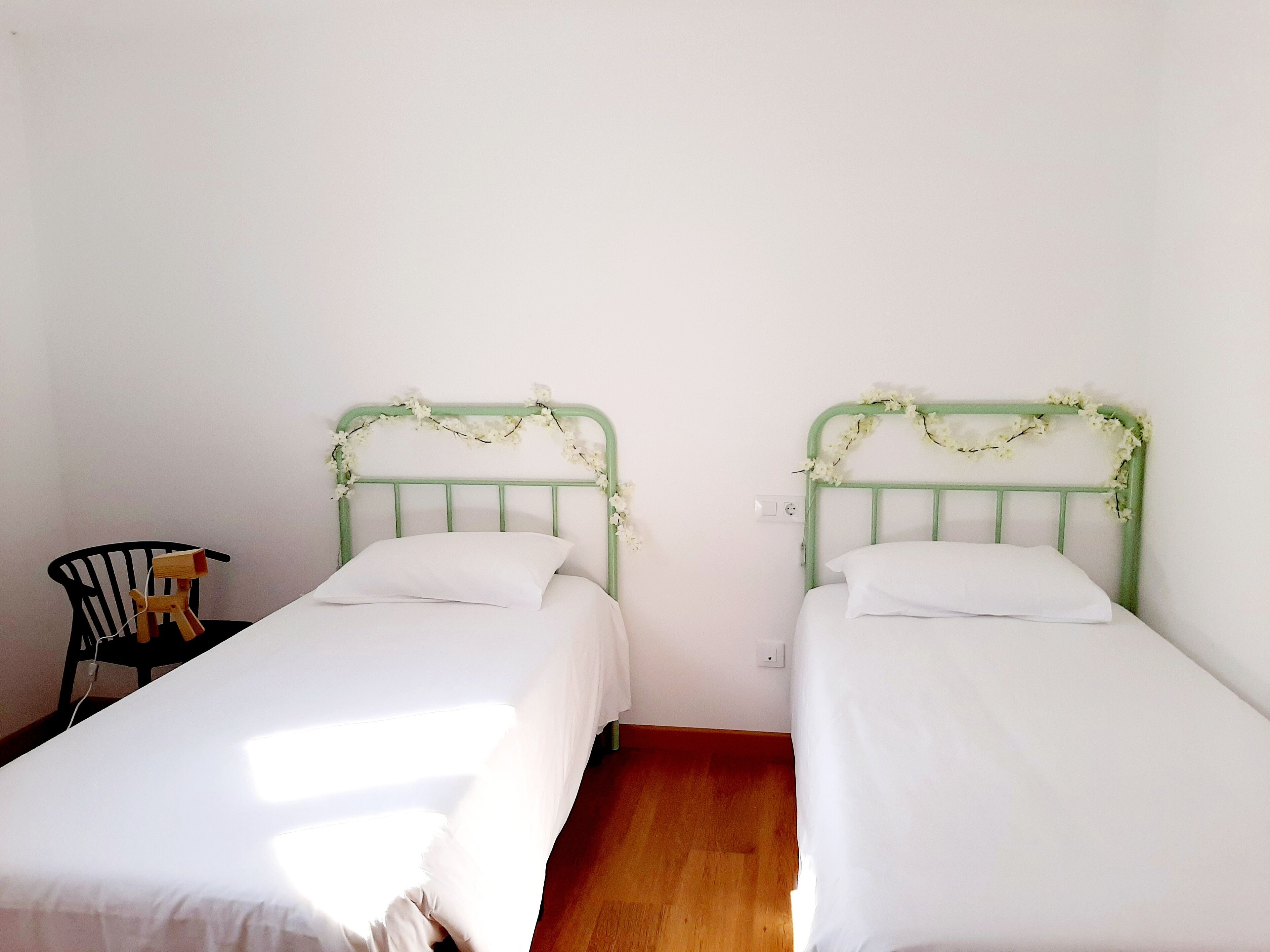 Dos camas individuales con sábanas blancas, una silla de madera y un cabecero de metal verde.