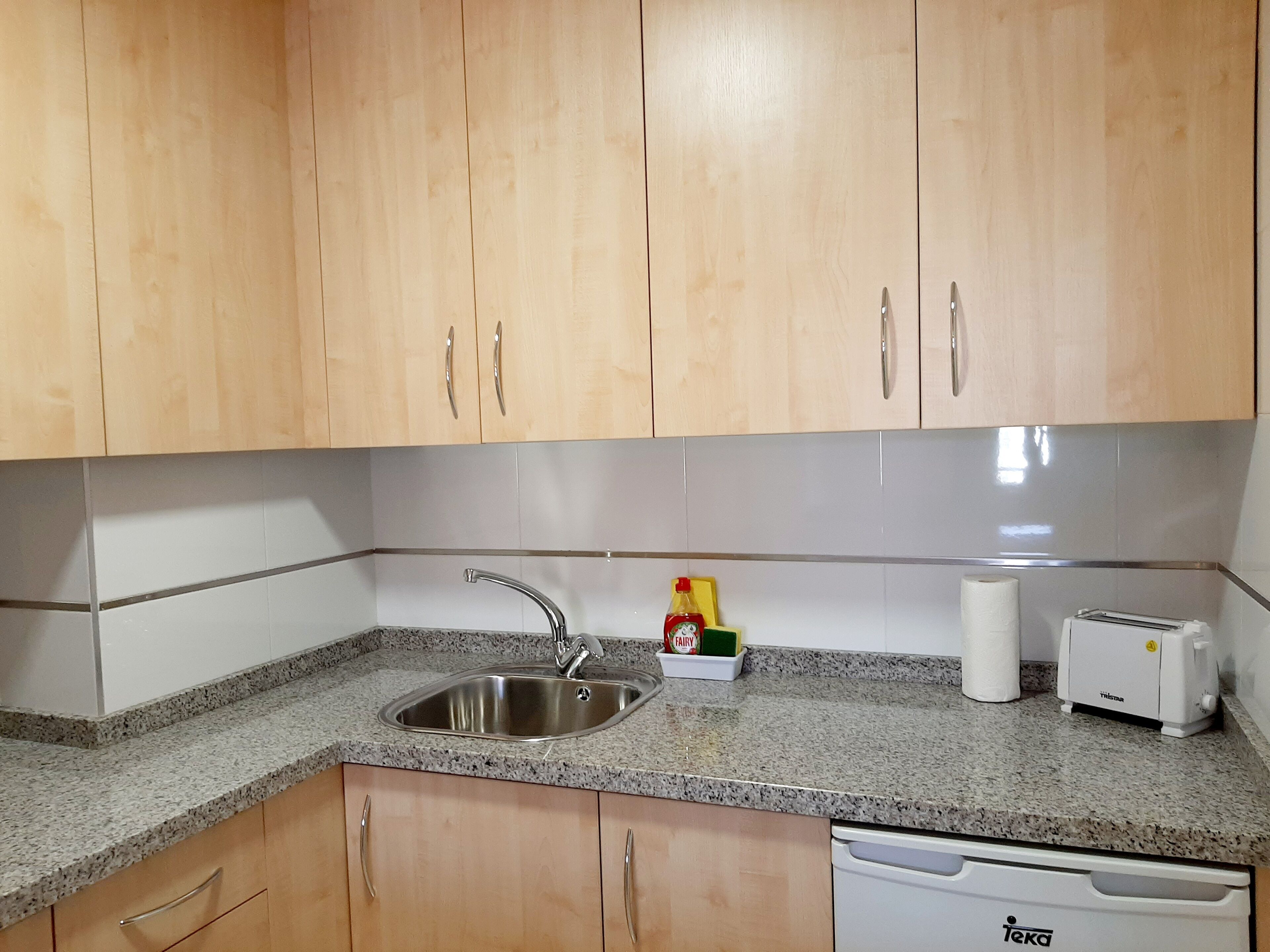 A kitchen with wooden cabinets, a stainless steel sink, a dish soap bottle, a paper towel dispenser, and a white dishwasher.