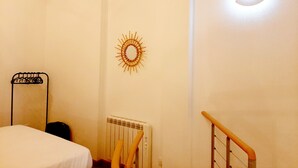 Habitación de hotel con cama, radiador, silla de madera, espejo adosado a la pared y un objeto decorativo con forma de sol.