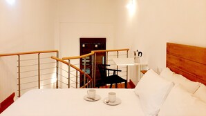 Habitación de hotel con cama, una mesita con taza y platillo, una silla y una escalera con barandilla.