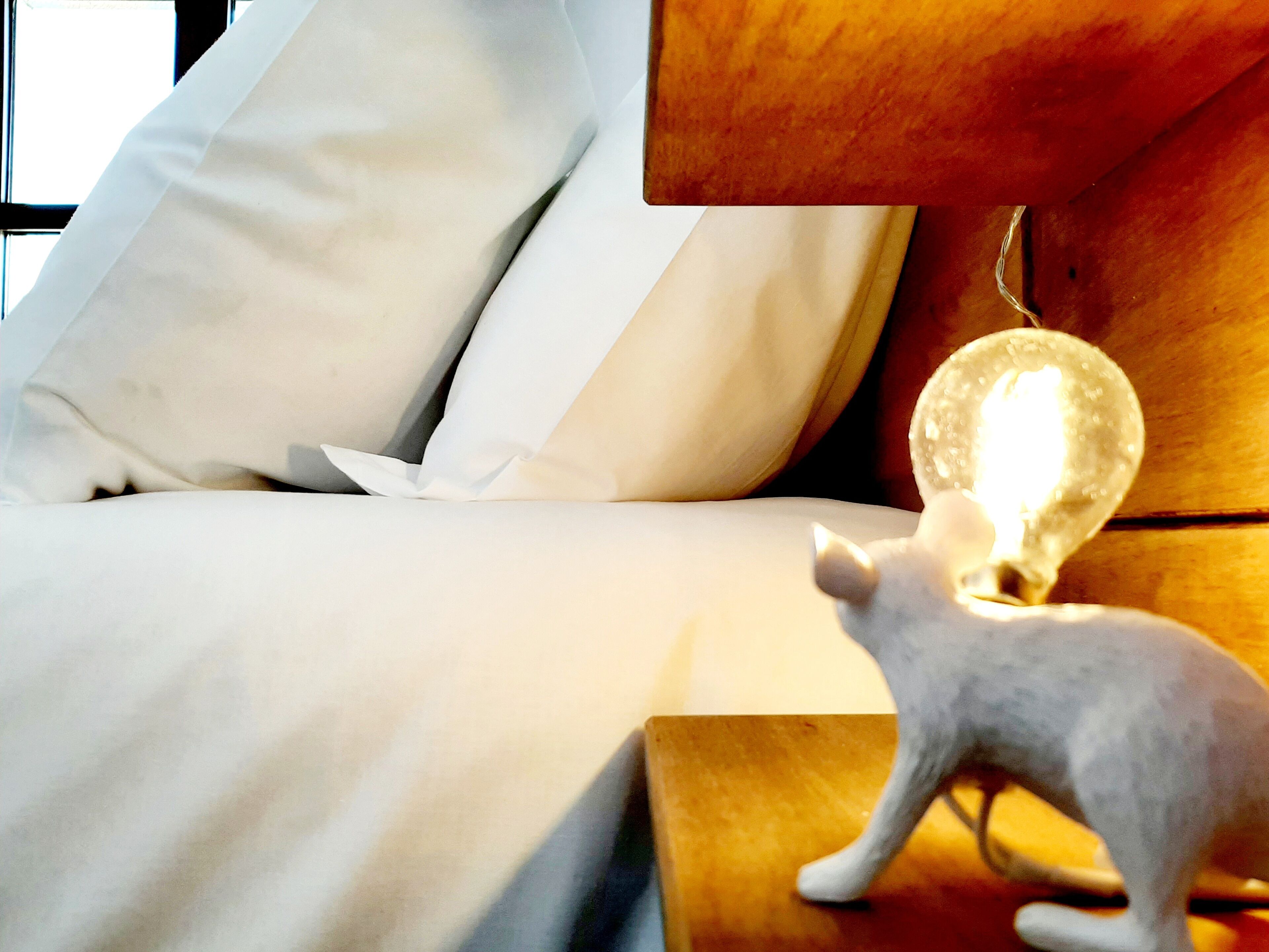 Un lit avec du linge de lit blanc et une petite statuette de chat blanc posée sur une table de chevet en bois, à côté d’une ampoule allumée.