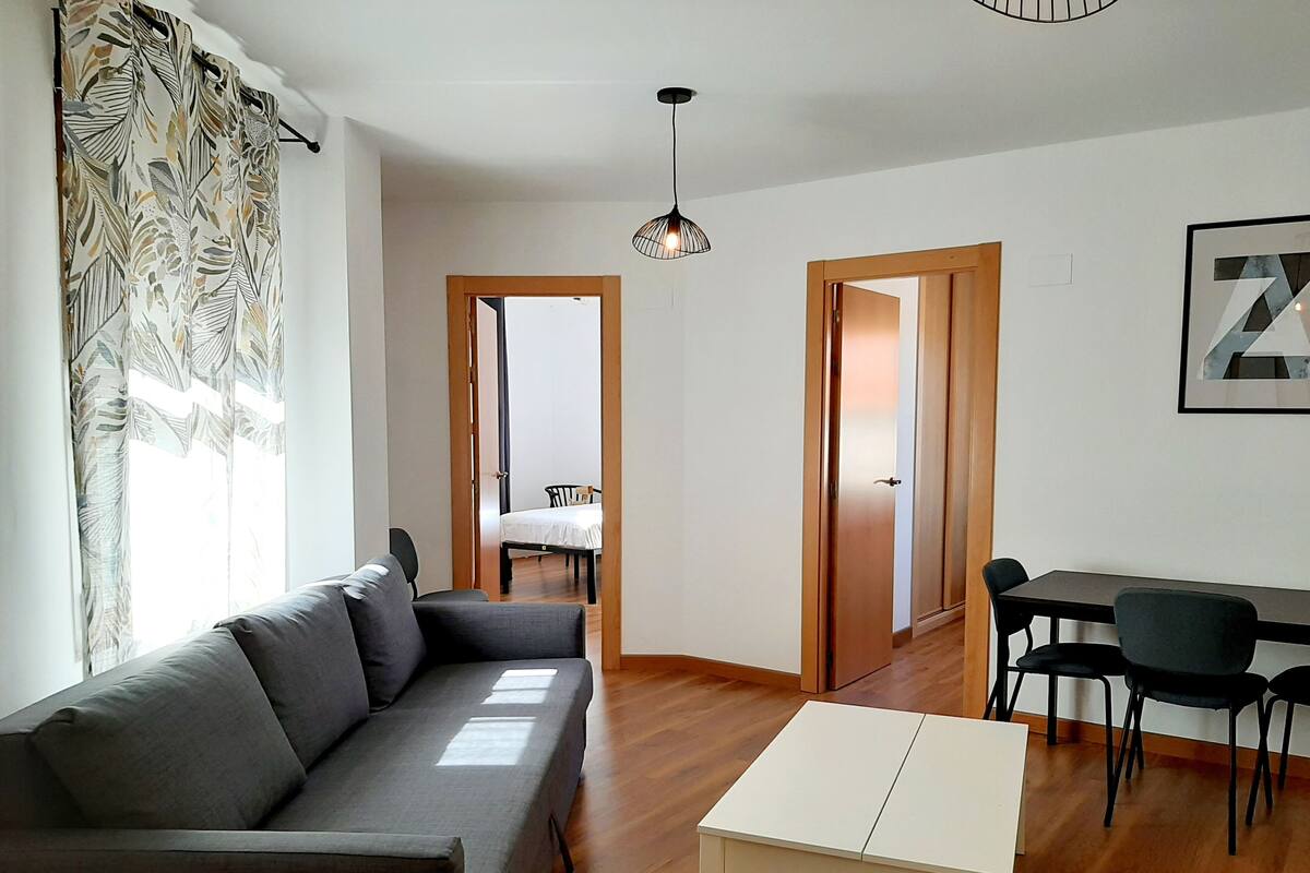 A modern living room with a sofa, a small table, and a dining area with chairs. There is a door leading to another room and a ceiling light fixture.